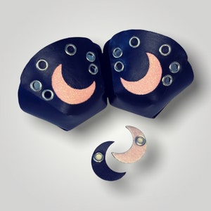 Puede incluir: Protectores de puntera de patines de color azul marino y accesorios a juego con forma de luna. Los protectores de puntera presentan un diseño de media luna rosa brillante y ojales plateados. Los accesorios incluyen un par de pendientes con forma de luna y un broche con forma de luna.