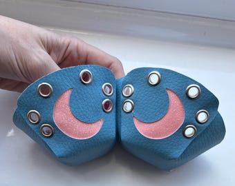 May include: Two blue leather wrist cuffs with pink glitter crescent moon cutouts and silver grommets. The cuffs are held up by a hand. The cuffs are a unique accessory.
