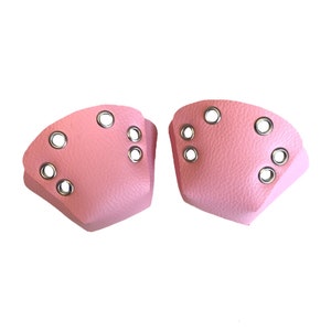 Könnte beinhalten: Zwei rosa Zehenkappen für Rollschuhe mit silbernen Ösen. Die Kappen bestehen aus einem strukturierten Material und sind so geformt, dass sie an die Vorderseite von Rollschuhen passen. Jede Kappe hat mehrere Ösen zur Befestigung an den Rollschuhen.