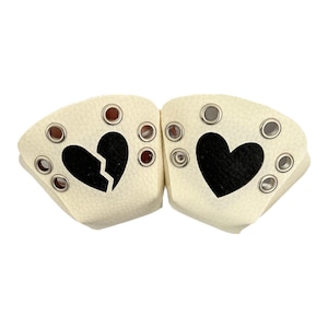 Op de afbeelding: Twee crèmekleurige schoenaccessoires met een zwart hartontwerp. Eén hart is gebroken, de andere is heel. Elk stuk heeft meerdere zilveren oogjes en bruine klinknagels.