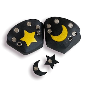 Puede incluir: Brazaletes de cuero negro con diseños de estrellas y lunas amarillas. Cada brazalete tiene ojales plateados. También se incluyen dos colgantes más pequeños a juego con forma de estrella y luna. Un accesorio con temática celestial.
