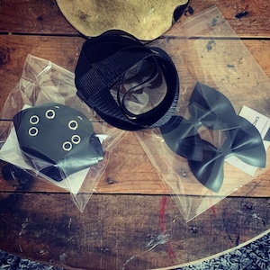 May include: A selection of black accessories, including a strap, a bow, and a piece with eyelets. The items are packaged in clear plastic and arranged on a wooden surface. The bow has a satin finish.
