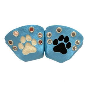 Peut inclure: Deux protège-pattes pour animaux de compagnie en cuir bleu clair. L'un a une empreinte de patte crème, l'autre une empreinte de patte noire. Chacun a plusieurs œillets argentés et œillets rouges.