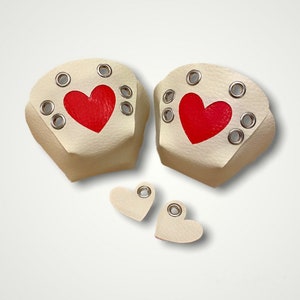 May include: Cream-coloured leather accessories with red heart accents. The set includes two larger pieces with silver grommets and a heart-shaped cutout, plus two smaller heart-shaped pieces, also with grommets. The design is simple and romantic.