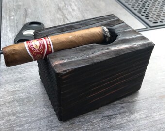 May include: A black wooden cigar holder with a cigar resting in the slot. The cigar is brown and has a red and gold band that says "Romeo y Julieta" and "Reserva" on it.