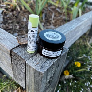 May include: A lip balm and a jar of salve are displayed on a weathered wooden surface. The lip balm is white with green accents and the word "HONEY" is visible. The jar is dark brown with a white label that reads "Herbal 'Triple Antibiotic' Salve."