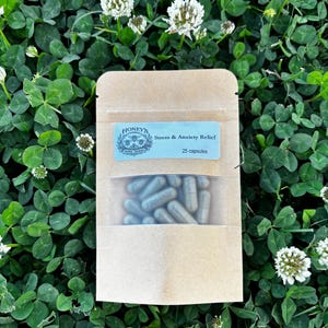May include: A kraft paper pouch with a clear window, containing blue-green capsules. The pouch has a blue label that reads "Honey's Stress & Anxiety Relief" with a floral design and "25 capsules". The background is a field of green clover and white flowers.