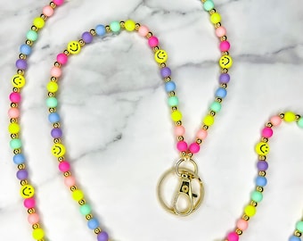 May include: A colorful beaded lanyard with a gold clasp and smiley face charms. The lanyard is made of pastel pink, yellow, green, blue, and purple beads.