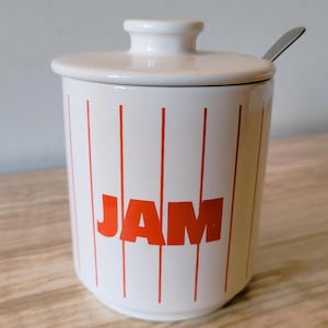 May include: White ceramic jam pot with a lid and a small spoon. The pot features vertical red stripes and the word "JAM" in red. The pot is on a wooden surface.