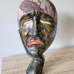 May include: A carved wooden mask featuring a black face with yellow distressed paint, bright red lips, and almond-shaped eye cutouts. The forehead area is covered in a light-coloured fabric with a dark brown geometric batik-style pattern.