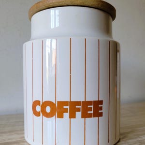 May include: White ceramic coffee jar with a wooden lid. The jar features vertical orange stripes and the word "COFFEE" in orange block letters. A kitchen storage container.