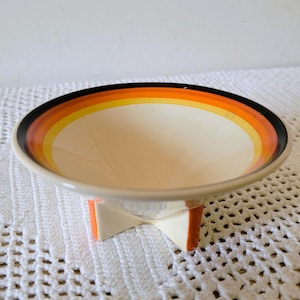 May include: A ceramic bowl with a cream-coloured interior and a wide, flared rim featuring concentric bands of orange, yellow, and black. The bowl is supported by a cross-shaped base in a matching cream colour with orange accents on the sides.