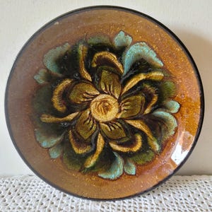 May include: A round ceramic plate featuring a central floral motif with layered petals in shades of deep brown, olive green, and turquoise. The background of the plate is a speckled amber-brown glaze with a dark rim and a glossy finish.