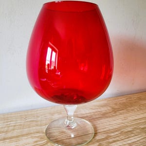 May include: A large, bulbous red glass vessel shaped like a brandy snifter, featuring a wide, rounded bowl that tapers towards the top opening. The vessel is supported by a short, clear glass stem and a circular, transparent glass base.