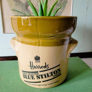 May include: A beige and olive green ceramic crock with two side handles. The front features black text reading Harrods Finest Selected Blue Stilton Cheese and Harrods Limited Knightsbridge London England, with a small crest above the text.