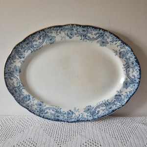 May include: An oval ceramic serving platter featuring a white centre and a wide border decorated with a dark blue floral and scroll pattern. The rim of the platter has a scalloped edge and is finished with a thin gold trim along the outer perimeter.