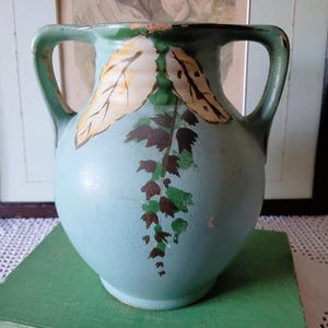 May include: A light blue ceramic vase with two handles, featuring a painted design of two large cream-coloured leaves at the top and a trailing vine with dark brown and green leaves extending downward towards the centre of the rounded body.