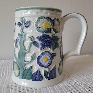 May include: A ceramic mug with a cream-coloured base featuring a raised, embossed floral pattern. The design includes blue and green leaves, vines, and flowers with yellow centres. A geometric border pattern encircles the top rim of the mug.