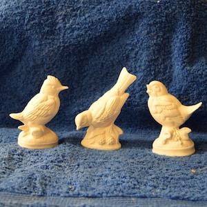 May include: Three white ceramic bird figurines, each perched on a base with a leafy design. The birds are in various poses, with one looking to the side, one looking down, and one looking forward.