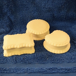 May include: Three ceramic boxes in different shapes, a rectangle, a circle, and a heart, all with a white glaze and decorative trim. The boxes are sitting on a blue towel.