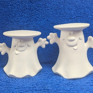 May include: Two white ceramic ghost candle holders with a whimsical design. Each ghost has a wide smile and outstretched arms. The top of each ghost is a flat surface for holding a candle.