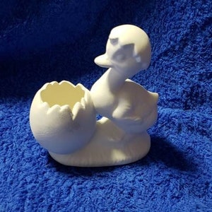 May include: White ceramic duck figurine holding a cracked egg. The duck is standing on a base and has a detailed beak and eyes. The egg is open and has a small hole in the top.