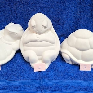 May include: Three white ceramic turtle figurines. The first turtle is lying on its back with its legs in the air. The second turtle is sitting upright with its arms crossed. The third turtle is walking with its head up. The turtles are all unpainted and ready to be decorated.