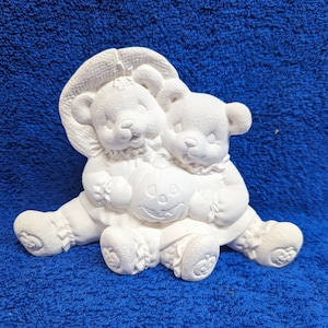 May include: Two white ceramic teddy bears dressed in Halloween costumes holding a jack-o'-lantern. The bears are sitting on a blue fuzzy surface.