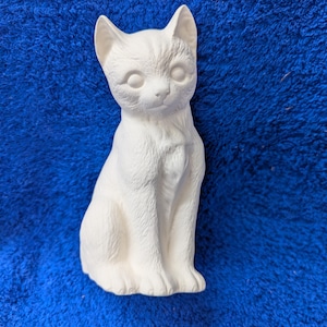 May include: White ceramic figurine of a cat sitting with its head tilted slightly to the side. The cat has a detailed face with closed eyes and a small nose.