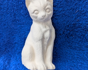 May include: White ceramic figurine of a cat sitting with its head tilted slightly to the side. The cat has a detailed face with closed eyes and a small nose.