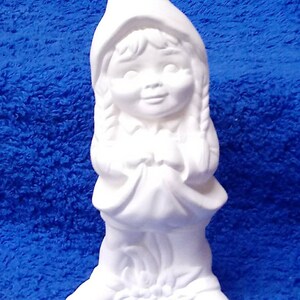 May include: White ceramic figurine of a girl wearing a hooded dress and holding her hands in front of her. The girl has long braids and a sweet expression on her face.