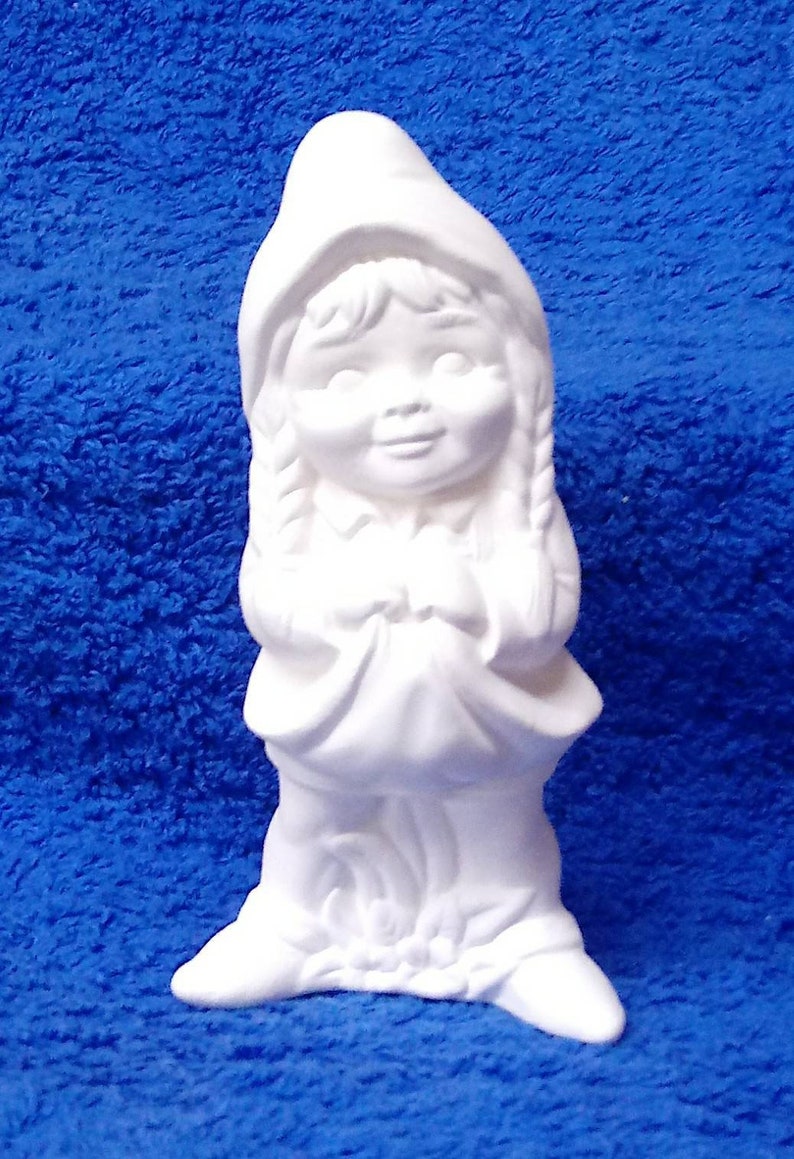 May include: White ceramic figurine of a girl wearing a hooded dress and holding her hands in front of her. The girl has long braids and a sweet expression on her face.