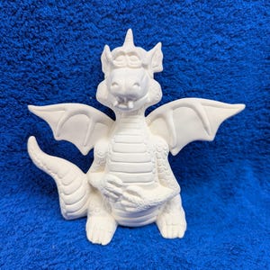 May include: A white ceramic dragon figurine with outstretched wings, a long tail, and a crown. The dragon is in a seated position, with its hands clasped together. The figurine has detailed scales and a friendly expression.