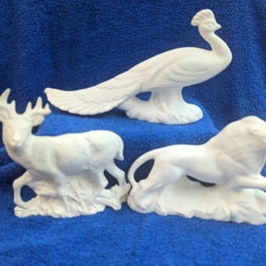 May include: Three white ceramic figurines of a deer, a peacock, and a lion. The deer is standing on a base with grass and leaves. The peacock is standing on a base with a rock. The lion is standing on a base with rocks.