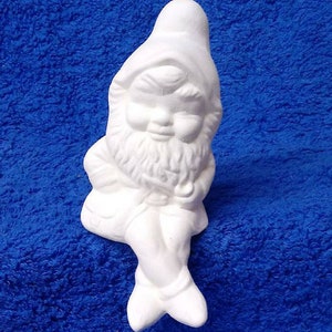 May include: White ceramic gnome figurine sitting with legs crossed and a beard. The gnome is wearing a pointed hat and has a closed-eye expression.