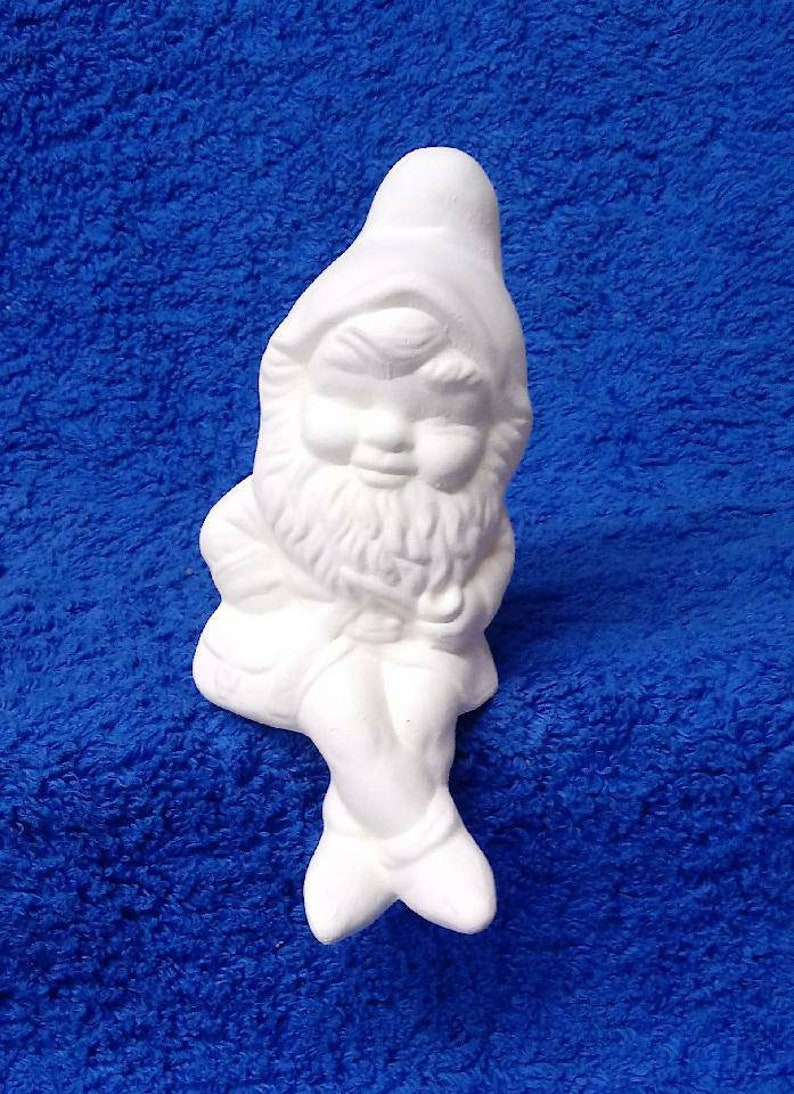 May include: White ceramic gnome figurine sitting with legs crossed and a beard. The gnome is wearing a pointed hat and has a closed-eye expression.