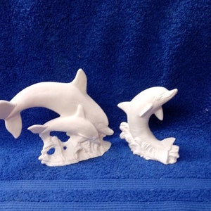 May include: Two white ceramic dolphin figurines. One dolphin is larger and has a smaller dolphin jumping out of its back. The smaller dolphin is jumping out of the water. Both dolphins are on a base that looks like water.