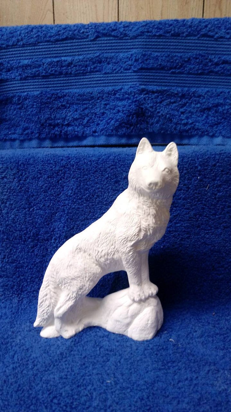 May include: White ceramic figurine of a wolf standing on a rock. The wolf is facing forward with its head tilted slightly up. The figurine is unpainted and ready to be decorated.