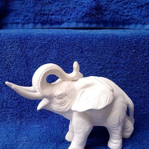 May include: White ceramic elephant figurine with its trunk raised and its ears spread out. The elephant is standing on a blue surface.