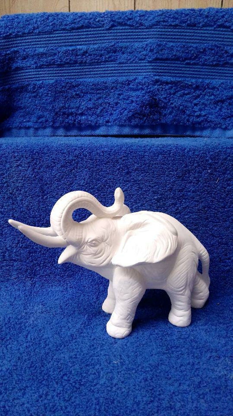 May include: White ceramic elephant figurine with its trunk raised and its ears spread out. The elephant is standing on a blue surface.