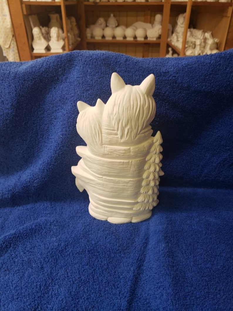 K&ouml;nnte beinhalten: Wei&szlig;e Keramikfigur von zwei B&auml;ren, die sich umarmen, einer sitzt auf dem anderen, mit einem Kiefernbaum dahinter. Die B&auml;ren sind in einem Design aus gestapelten Blockh&uuml;tten.
