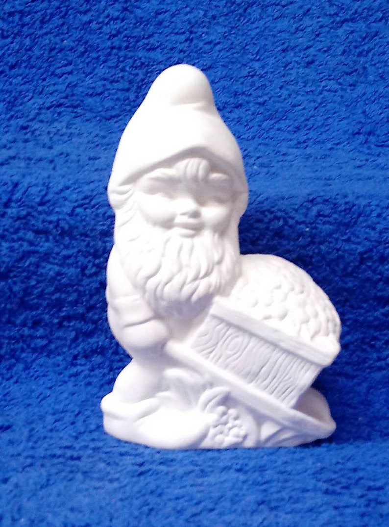 May include: White ceramic gnome figurine holding a wheelbarrow filled with small round objects. The gnome has a beard and a pointed hat.