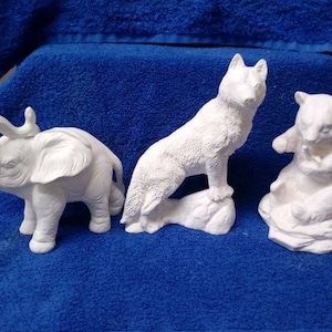 May include: Three white ceramic figurines of an elephant, wolf, and bear. The elephant has its trunk raised in the air. The wolf is standing on a rock. The bear is sitting on a rock.