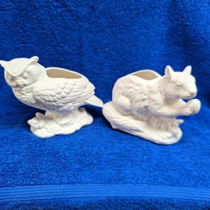 May include: Two white ceramic planters shaped like an owl and a squirrel. The owl planter has a detailed design with feathers and a beak. The squirrel planter has a detailed design with fur and a bushy tail. Both planters have a small hole in the top for planting.