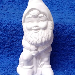 May include: White ceramic gnome figurine with a beard and a hat. The gnome is holding a rake in its hands and is standing on a base.