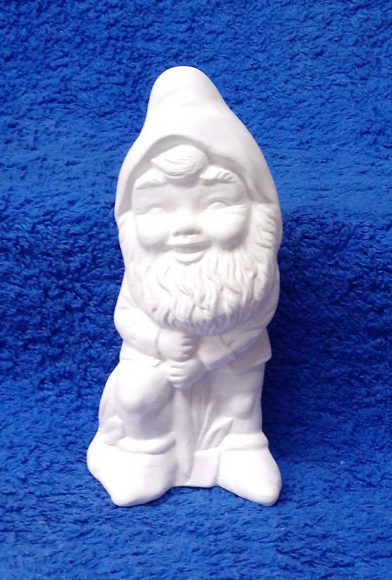 May include: White ceramic gnome figurine with a beard and a hat. The gnome is holding a rake in its hands and is standing on a base.