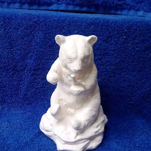 May include: White ceramic figurine of a bear sitting on a rock. The bear is holding a small object in its paws.