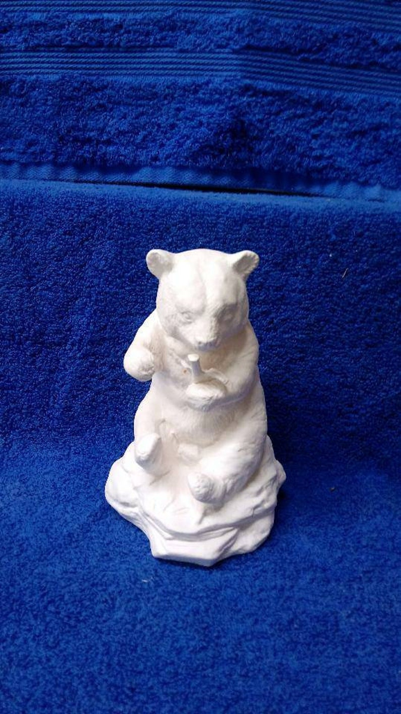 May include: White ceramic figurine of a bear sitting on a rock. The bear is holding a small object in its paws.