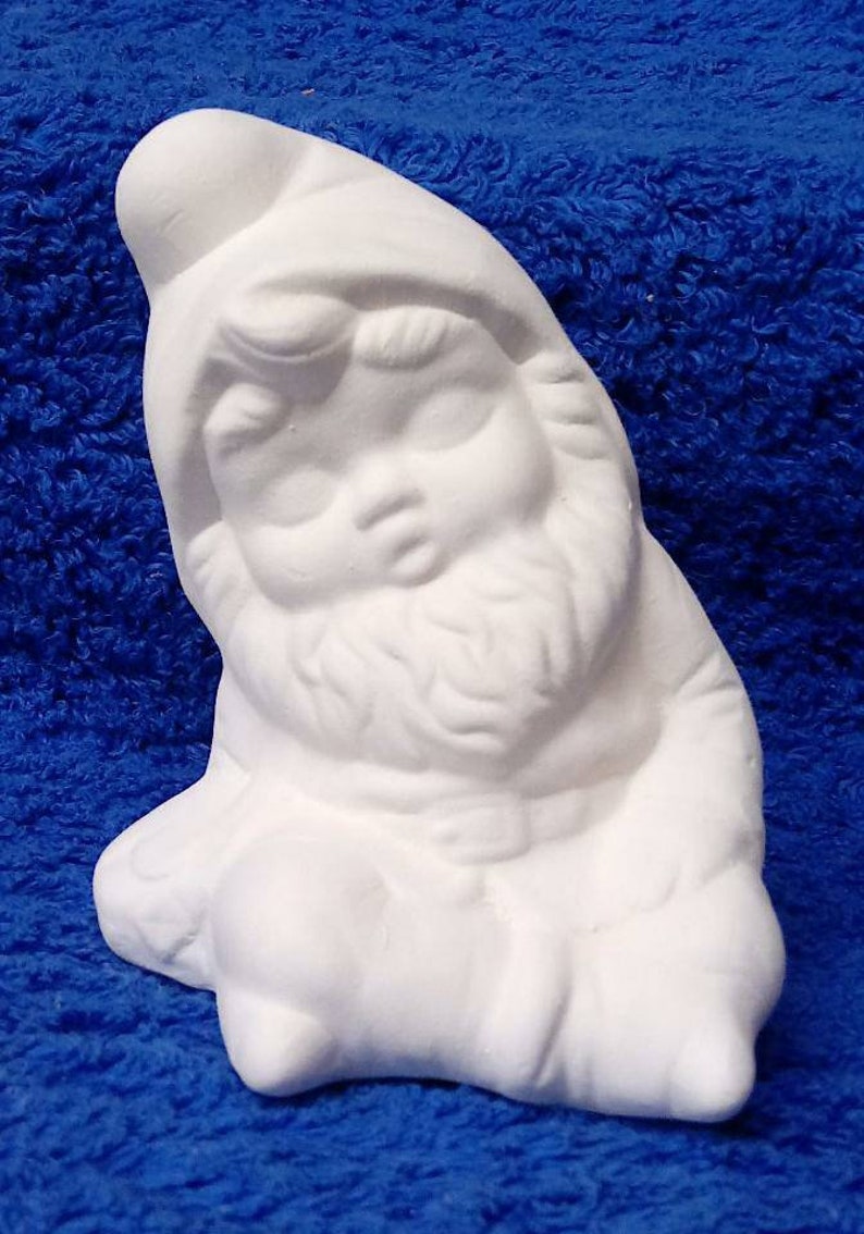 May include: White ceramic gnome figurine with a rounded hat, closed eyes, and a beard. The gnome is sitting with its legs crossed and its hands resting on its lap.