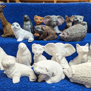 May include: A collection of 14 unpainted ceramic animal figurines, including a giraffe, elephant, penguin, seal, cat, bear, and hedgehog. The figurines are arranged on a blue surface.